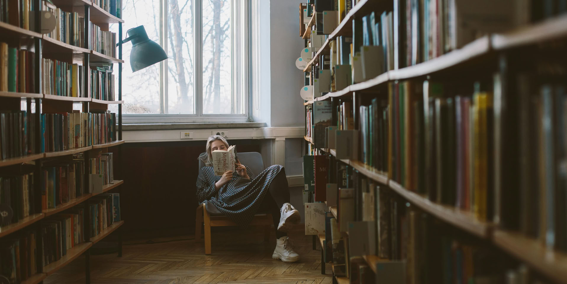 Woman enjoying a quiet read in a sunlit library corner with rows of bookshelves.