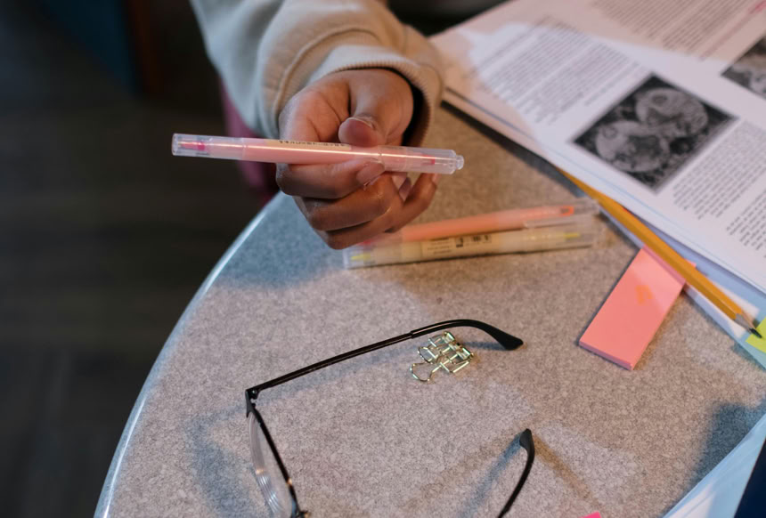 Overhead view of a person studying with books and holding highlighters on a table.